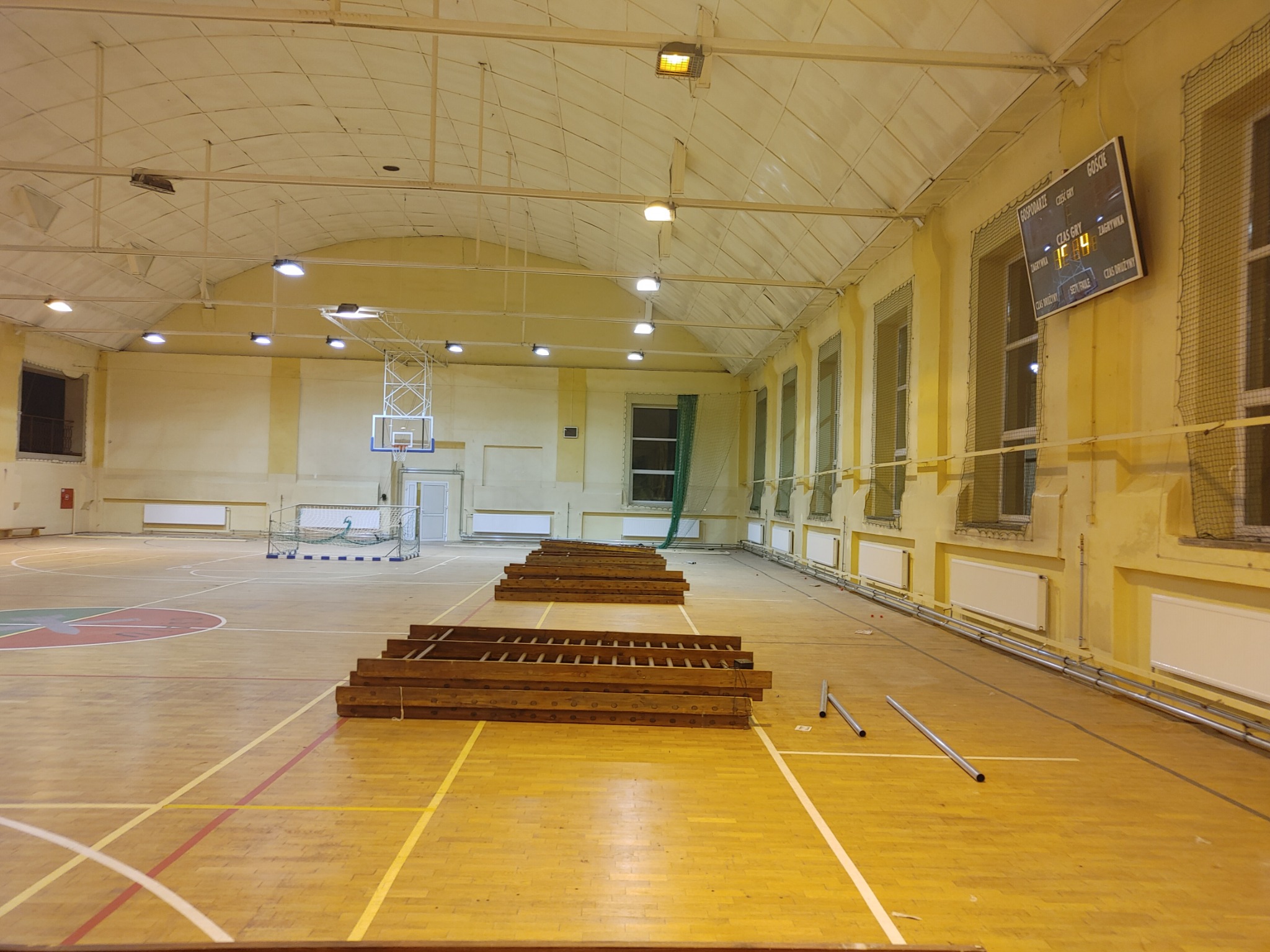 Sala gimnastyczna z drewnianą podłogą, na której leżą zdemontowane grzejniki i drabiny, przygotowana do prac instalacyjnych.