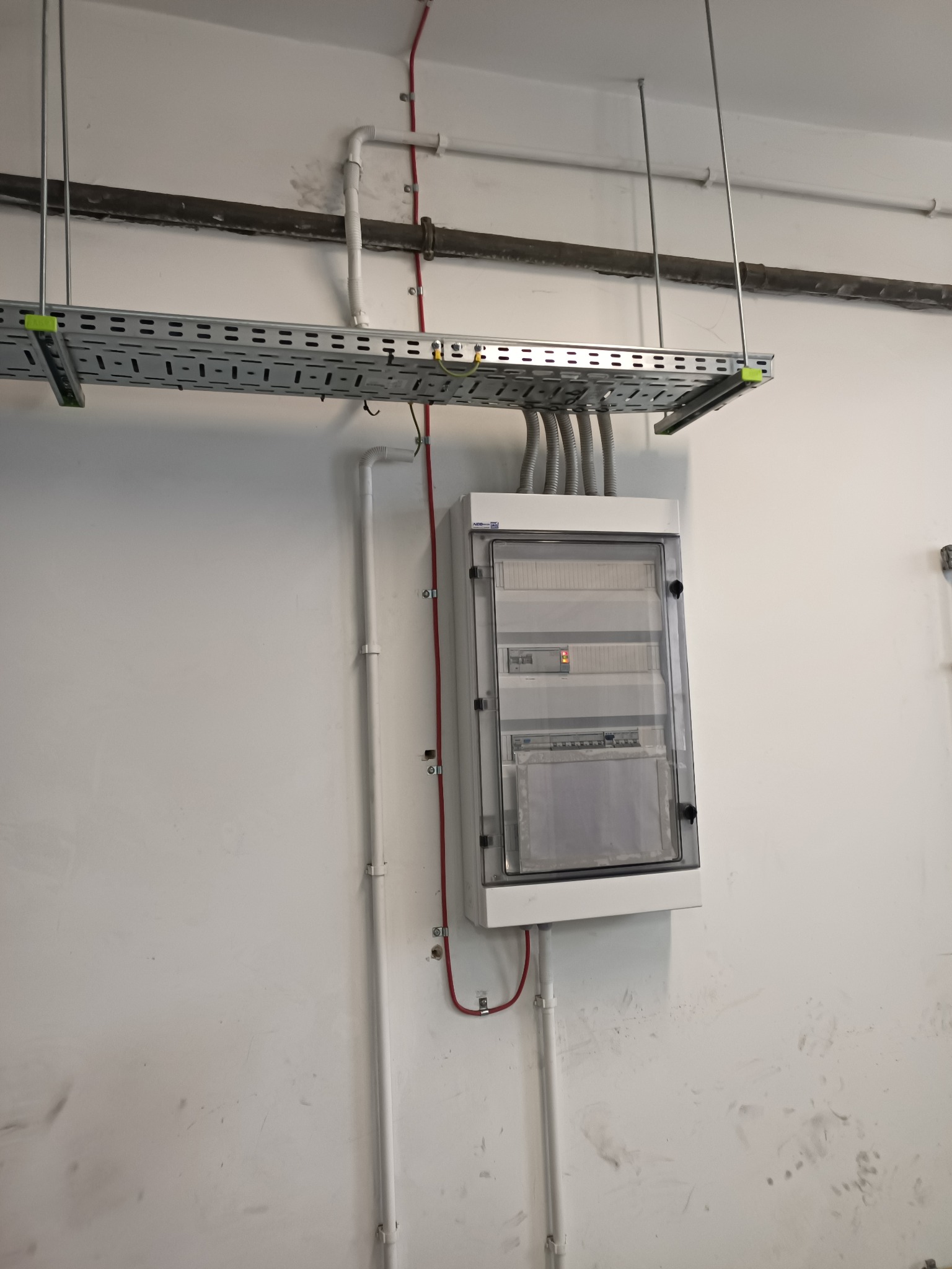 Szara szafa elektryczna z przezroczystymi drzwiami, zamontowana na białej ścianie, z widocznymi przewodami w metalowej korycie podwieszonym pod sufitem i rurami instalacyjnymi.