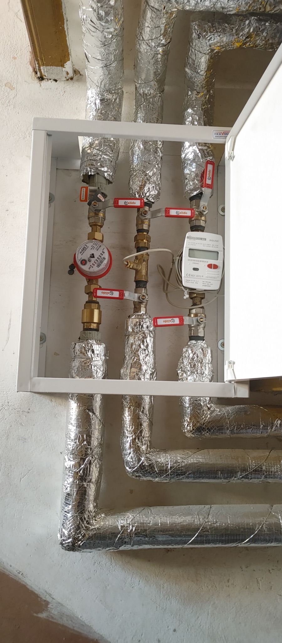 Kominacja rur grzewczych z zaworami Calido i licznikiem ciepła, zabezpieczonych folią izolacyjną wewnątrz białej szafki na tle ściany.