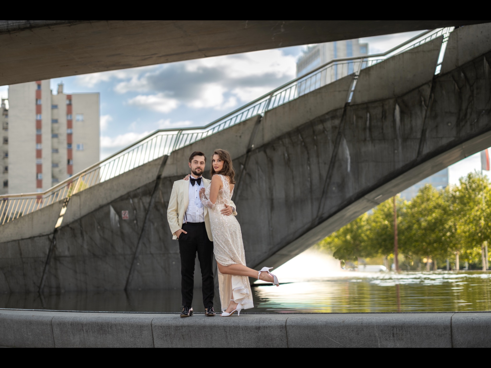 Elegancka para narzeczeńska pozuje pod betonowym wiaduktem z miejskim krajobrazem w tle, odbicie w wodzie, on w kremowym smokingu z muszką, ona w cekinowej sukni z koronkowymi rękawami i białych...
