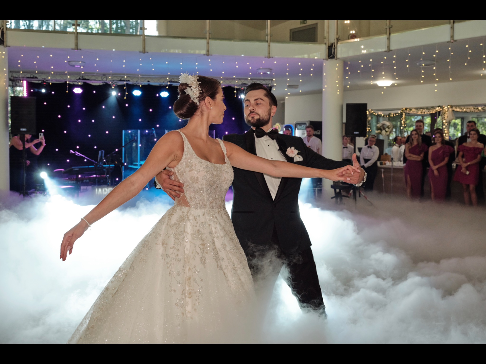 Para młoda w tańcu podczas wesela, otoczona sztuczną mgłą, w tle zespół muzyczny i goście.