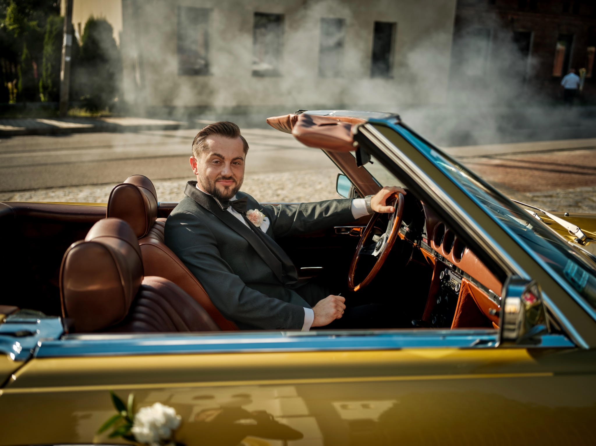 Elegancki mężczyzna w garniturze i muszce siedzi za kierownicą zabytkowego, żółtego kabrioletu z brązowym wnętrzem, udekorowanego białymi kwiatami, w tle budynek i lekka mgła