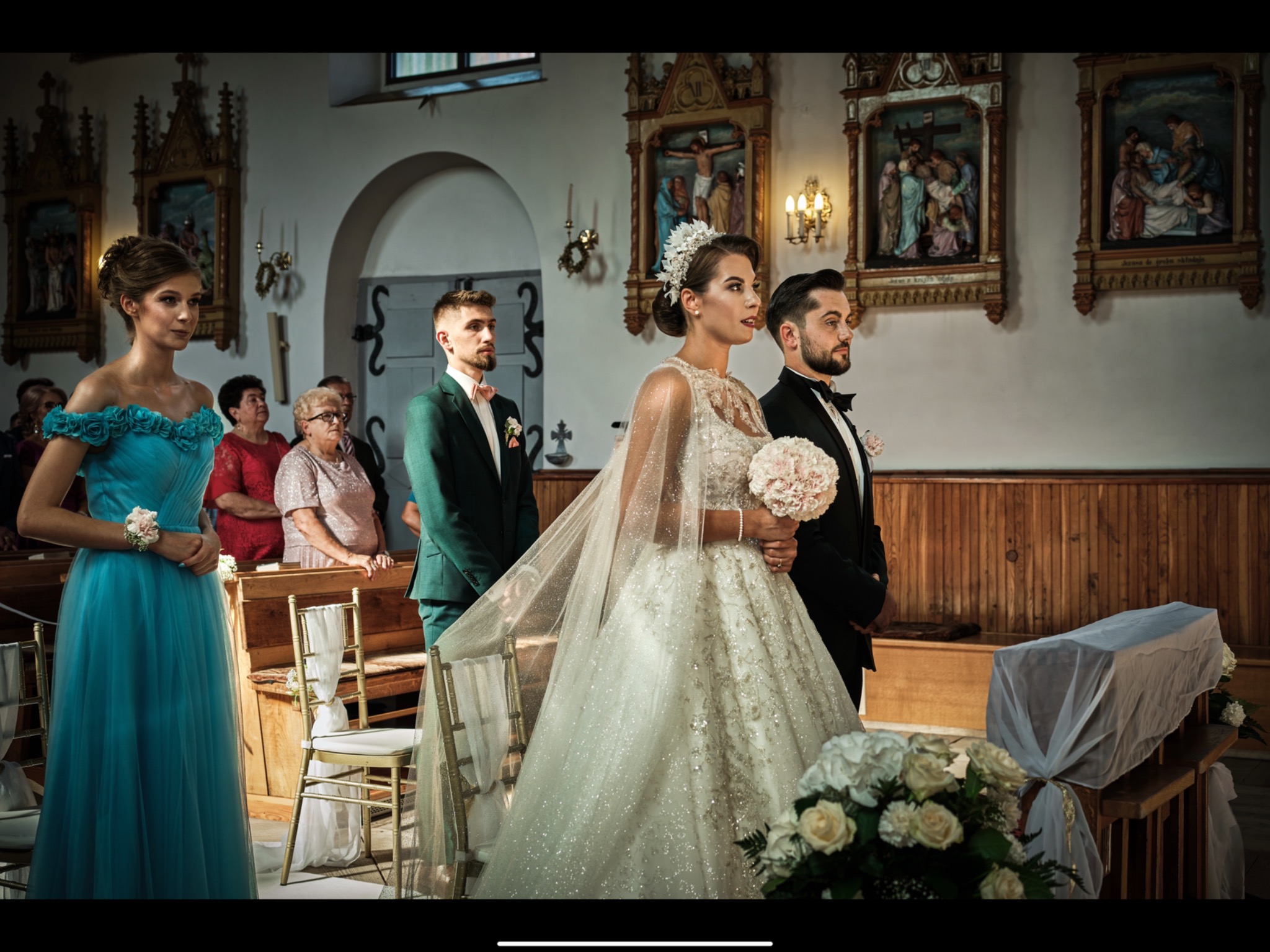 Para młoda podczas ceremonii ślubnej w kościele, panna młoda w bogato zdobionej sukni z welonem, pan młody w czarnym smokingu, druhna w turkusowej sukni, w tle obrazy religijne.