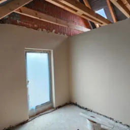 Poddasze po tynkowaniu maszynowym, widoczne drewniane krokwie, fragment muru z cegły, okno zabezpieczone folią i wiadro z narzędziami na podłodze.