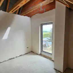 Poddasze z oknem po nałożeniu tynków, widoczne drewniane elementy konstrukcji dachu i fragment ściany z cegły.