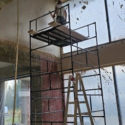 GP INSTAL Grzegorz Pricop - Tynkarz na rusztowaniu, wyrównuje ścianę w surowym wnętrzu z dużymi oknami zabezpieczonymi folią, widoczne instalacje pod sufitem.