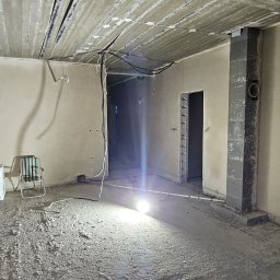 GP INSTAL Grzegorz Pricop - Surowe wnętrze w trakcie prac wykończeniowych: ściany pokryte świeżym tynkiem, betonowy sufit z widocznymi kablami, posadzka jastrychowa w trakcie schnięcia, oświetlone halogenem, z krzesłami...