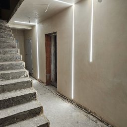 GP INSTAL Grzegorz Pricop - Surowy stan klatki schodowej z betonowymi schodami, świeżo otynkowanymi ścianami i nowoczesnym oświetleniem liniowym LED w ciepłej barwie, z widocznym niedokończonym otworem drzwiowym i wiszącą...