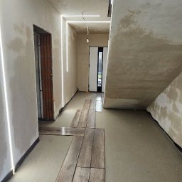 GP INSTAL Grzegorz Pricop - Surowy korytarz w trakcie wykańczania z niedokończonymi ścianami, posadzką jastrychową i rozłożonymi panelami podłogowymi imitującymi drewno oraz liniowym oświetleniem LED na suficie i ścianach.