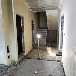 GP INSTAL Grzegorz Pricop - Wnętrze w trakcie prac wykończeniowych: świeżo otynkowane ściany, widoczne przewody, lampa budowlana oświetlająca schody w głębi pomieszczenia, rura wentylacyjna przy ścianie.