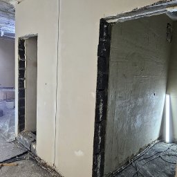 GP INSTAL Grzegorz Pricop - Surowe wnętrze w trakcie prac tynkarskich, widoczne świeżo otynkowane ściany w jasnym odcieniu, surowy betonowy strop, fragment instalacji elektrycznej na podłodze i nieotynkowane ościeżnice...