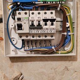 GP INSTAL Grzegorz Pricop - Rozdzielnica elektryczna z zamontowanymi bezpiecznikami IDEAL, okablowaniem oznaczonym opisami na przewodach, umieszczona na surowej ścianie z widoczną strukturą i fragmentami starego tynku.