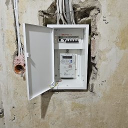 GP INSTAL Grzegorz Pricop - Otwarta, biała skrzynka elektryczna z bezpiecznikami i licznikiem, zamontowana w surowej ścianie z widocznymi kablami.