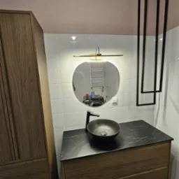 Nowoczesna łazienka z umywalką na szafce z drewnianymi frontami, okrągłym lustrem podświetlonym listwą LED i czarnymi, geometrycznymi elementami dekoracyjnymi na ścianie.