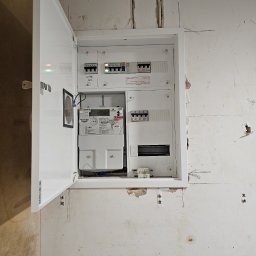 GP INSTAL Grzegorz Pricop - Otwarta biała skrzynka rozdzielcza z licznikiem i bezpiecznikami zamontowana na surowej, nieotynkowanej ścianie. Widoczne ślady po instalacji i rysunki na ścianie.