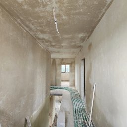 GP INSTAL Grzegorz Pricop - Wnętrze w stanie surowym: ściany pokryte świeżym tynkiem, widoczne przewody elektryczne na suficie i rury instalacyjne na podłodze, przygotowane do dalszych prac wykończeniowych.