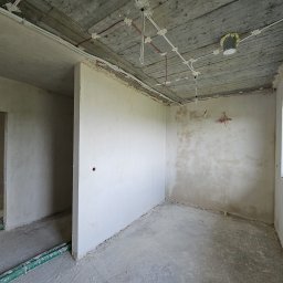 GP INSTAL Grzegorz Pricop - Surowy stan wnętrza z widocznymi przewodami elektrycznymi na suficie, częściowo otynkowane ściany, okno z widokiem na zieleń i fragment instalacji ogrzewania podłogowego.