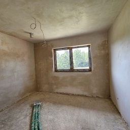 GP INSTAL Grzegorz Pricop - Surowe wnętrze pokoju z oknem i wstępnie nałożonym tynkiem na ścianach i suficie, widoczne wystające kable elektryczne i rury przy podłodze.