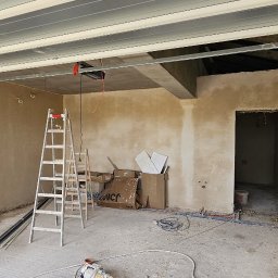 GP INSTAL Grzegorz Pricop - Wnętrze w stanie surowym, ściany pokryte świeżym tynkiem, widoczne dwie drabiny, rury instalacyjne na podłodze, okno z widokiem na zewnątrz i elementy konstrukcji sufitu.