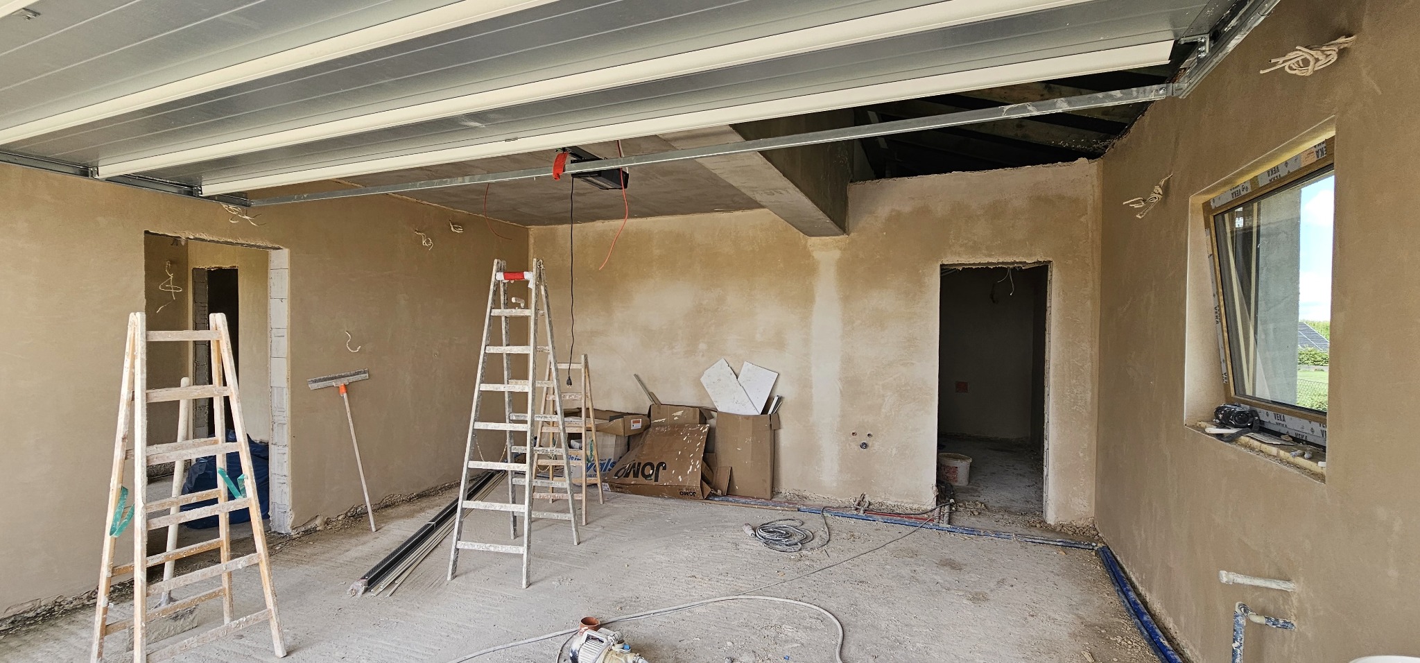 Wnętrze w stanie surowym w Rybniku po nałożeniu tynków, widoczne dwie drabiny, okno z parapetem, instalacje wystające ze ściany oraz sufit z metalową konstrukcją.