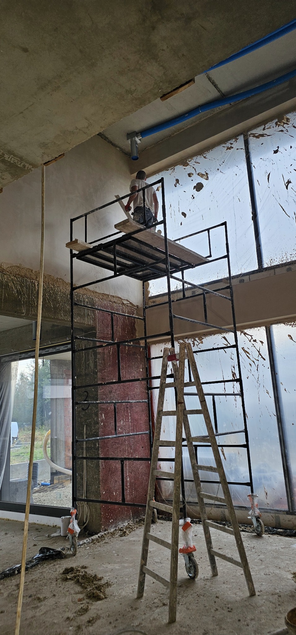Tynkarz na rusztowaniu, wyrównuje ścianę w surowym wnętrzu z dużymi oknami zabezpieczonymi folią, widoczne instalacje pod sufitem.