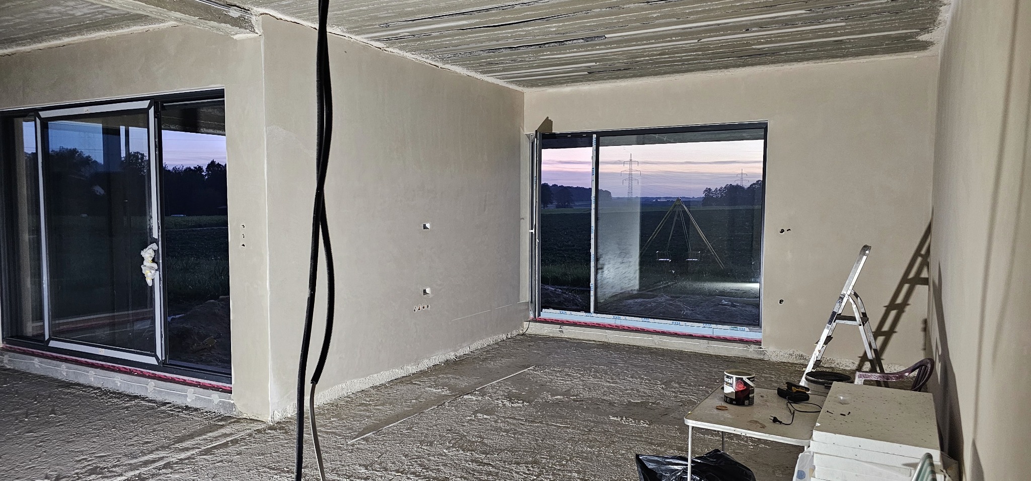 Surowe wnętrze w trakcie prac wykończeniowych: ściany po tynkowaniu maszynowym, posadzka jastrychowa w trakcie schnięcia, widoczne okna z widokiem na zewnątrz, drabina i narzędzia na stole.