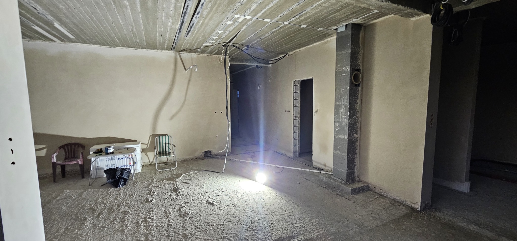 Surowe wnętrze w trakcie prac wykończeniowych: ściany pokryte świeżym tynkiem, betonowy sufit z widocznymi kablami, posadzka jastrychowa w trakcie schnięcia, oświetlone halogenem, z krzesłami...