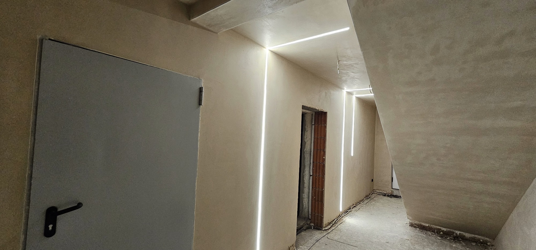 Korytarz w trakcie remontu z nałożonymi tynkami w odcieniach beżu i zamontowanym pionowym oświetleniem LED wzdłuż ścian. Widoczny fragment schodów i drzwi.