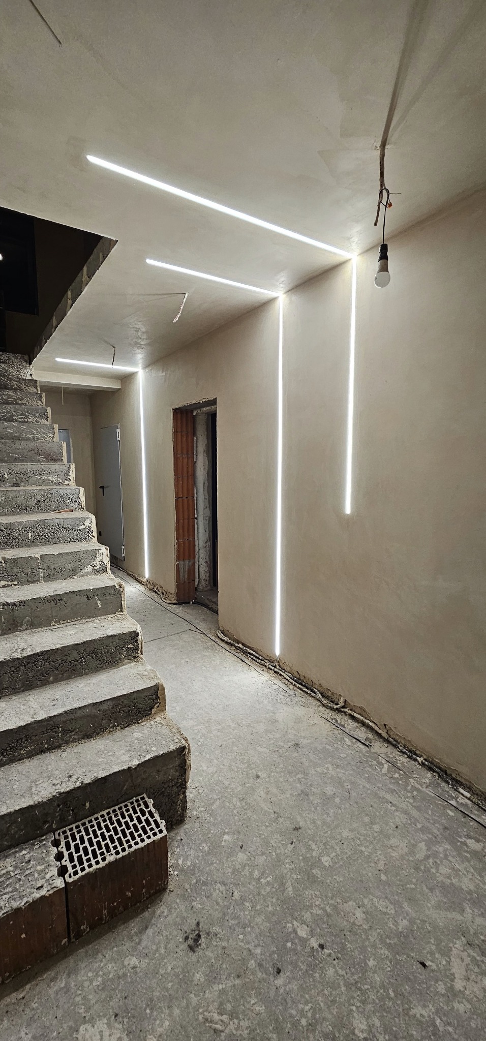Surowy stan klatki schodowej z betonowymi schodami, świeżo otynkowanymi ścianami i nowoczesnym oświetleniem liniowym LED w ciepłej barwie, z widocznym niedokończonym otworem drzwiowym i wiszącą...