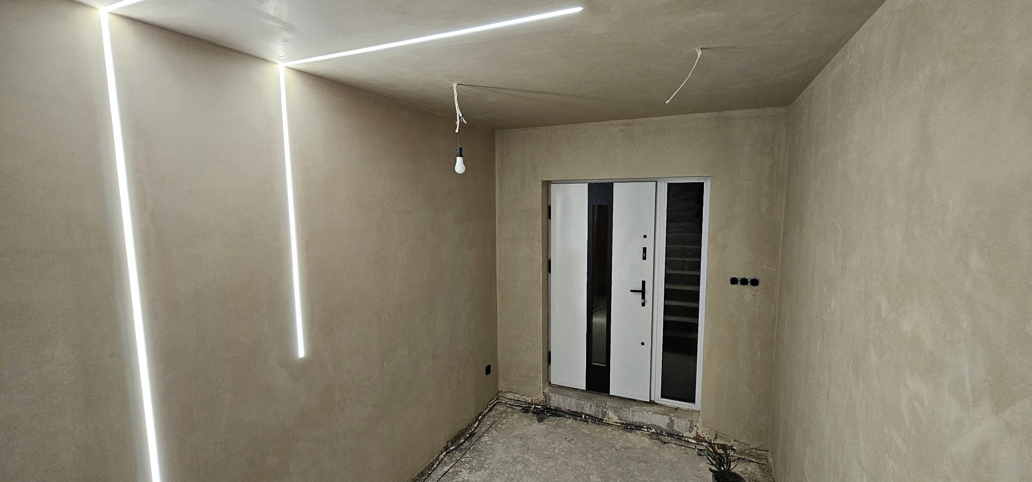 Wnętrze w trakcie wykańczania: tynki w odcieniach beżu, pionowe pasy oświetlenia LED na ścianie, widoczna instalacja elektryczna, surowa podłoga i biało-czarne drzwi wejściowe.