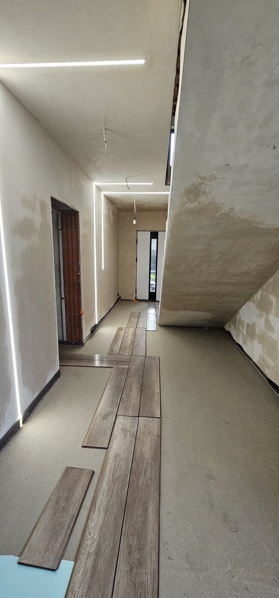 Surowy korytarz w trakcie wykańczania z niedokończonymi ścianami, posadzką jastrychową i rozłożonymi panelami podłogowymi imitującymi drewno oraz liniowym oświetleniem LED na suficie i ścianach.