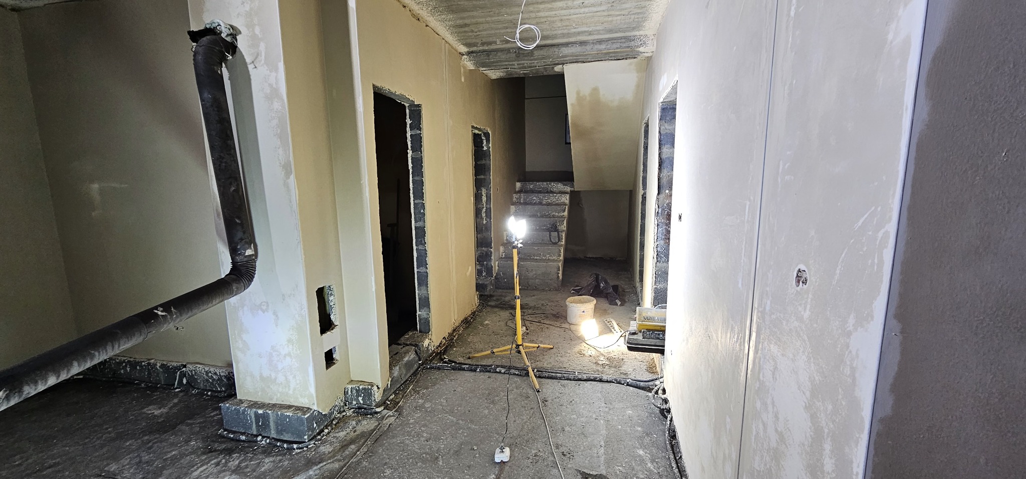 Wnętrze w trakcie prac wykończeniowych: świeżo otynkowane ściany, widoczne przewody, lampa budowlana oświetlająca schody w głębi pomieszczenia, rura wentylacyjna przy ścianie.