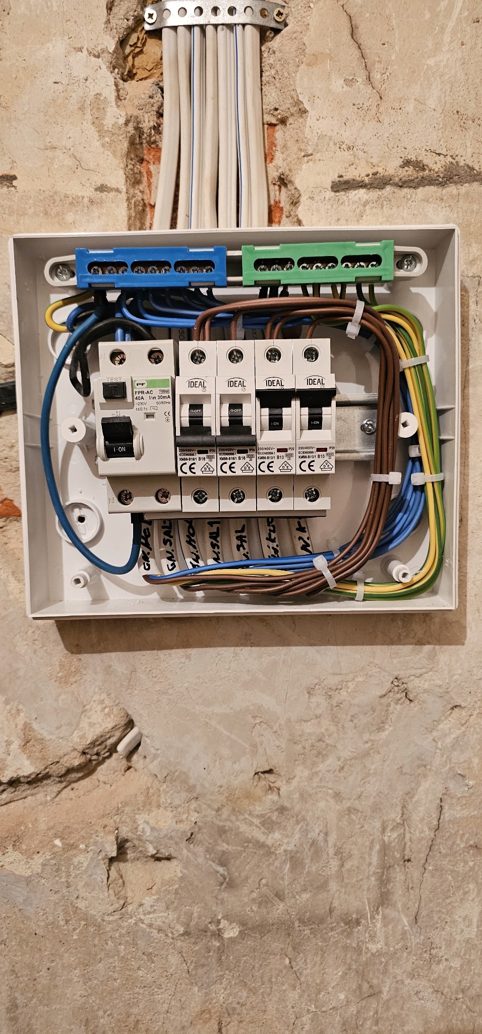 Rozdzielnica elektryczna z zamontowanymi bezpiecznikami IDEAL, okablowaniem oznaczonym opisami na przewodach, umieszczona na surowej ścianie z widoczną strukturą i fragmentami starego tynku.