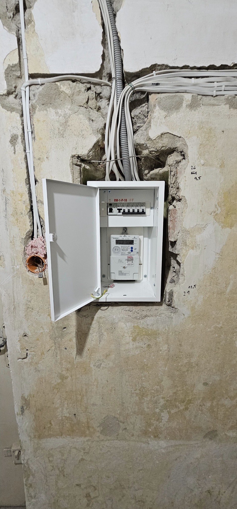 Otwarta, biała skrzynka elektryczna z bezpiecznikami i licznikiem, zamontowana w surowej ścianie z widocznymi kablami.