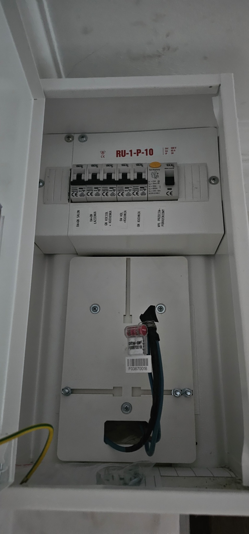 Wnętrze białej skrzynki elektrycznej z zamontowanymi bezpiecznikami i okablowaniem, widoczne oznaczenia obwodów: salon, łazienka, kocioł, kuchnia, hol, oraz zabezpieczenie przeciwporażeniowe.