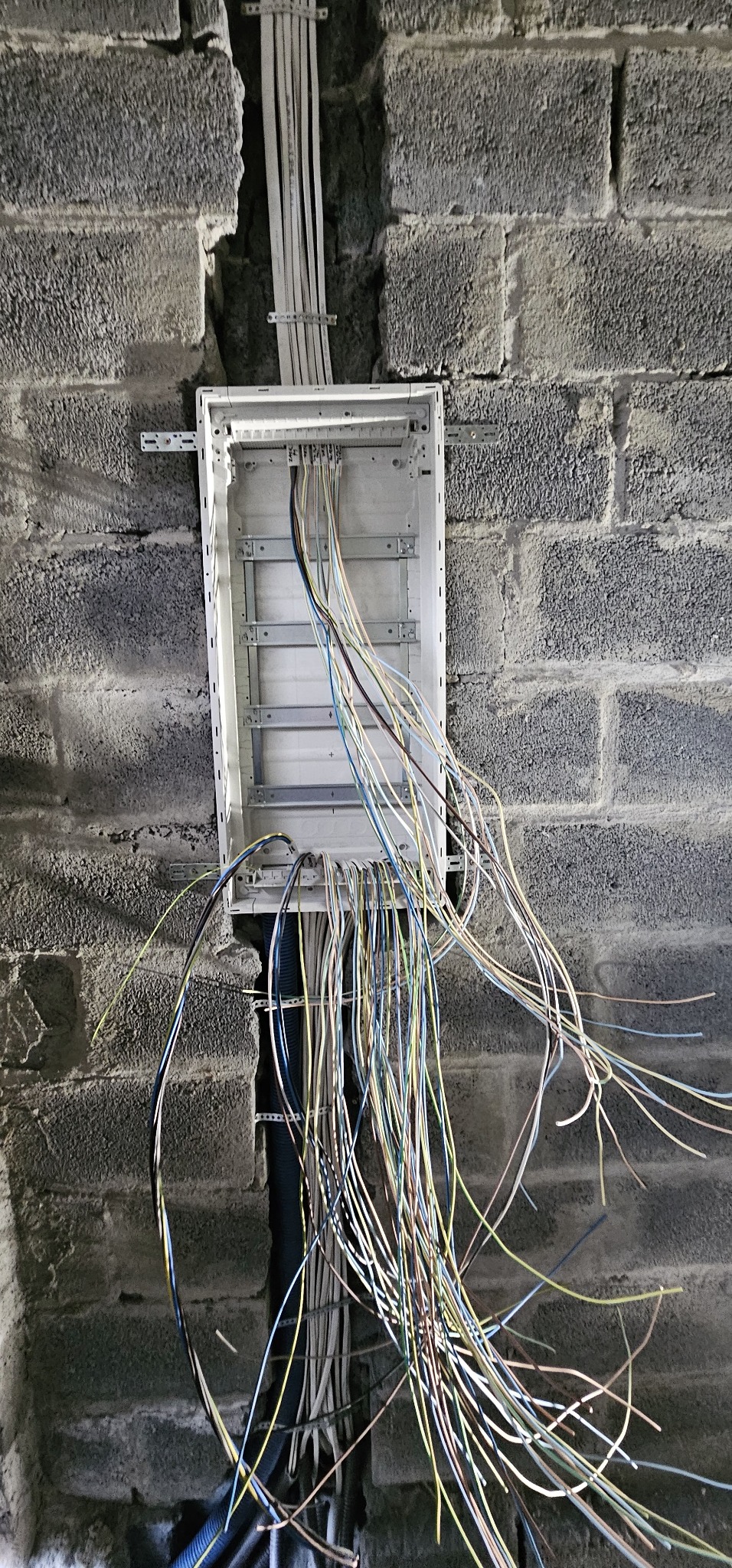 Rozdzielnica elektryczna zamontowana na szarej ścianie z bloczków betonowych, z widocznymi przewodami wychodzącymi z puszki i prowadzącymi w dół oraz w górę ściany.