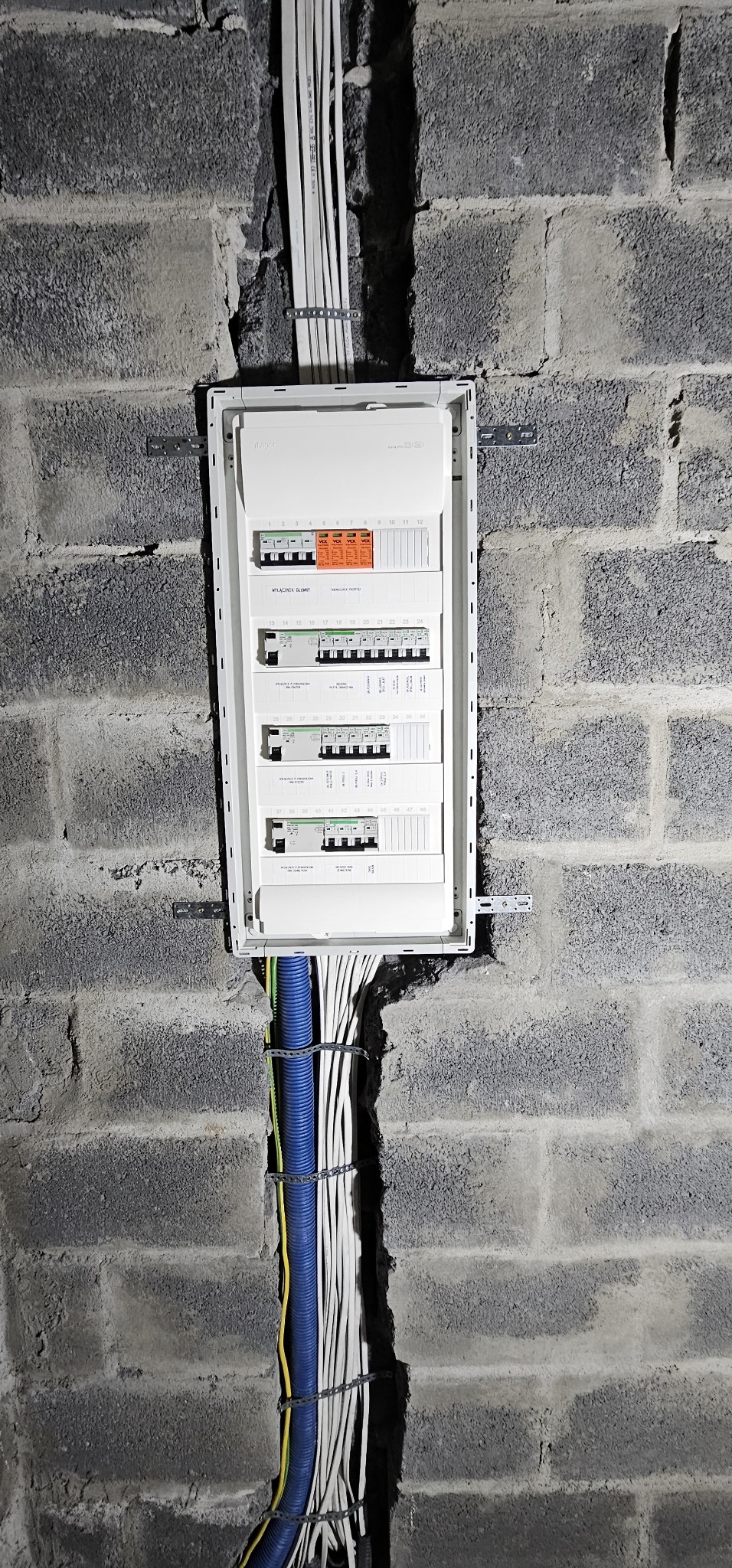 Rozdzielnica elektryczna zamontowana na szarej ścianie z betonu komórkowego, z widocznymi przewodami w peszlu i białymi kablami przymocowanymi metalowymi uchwytami.