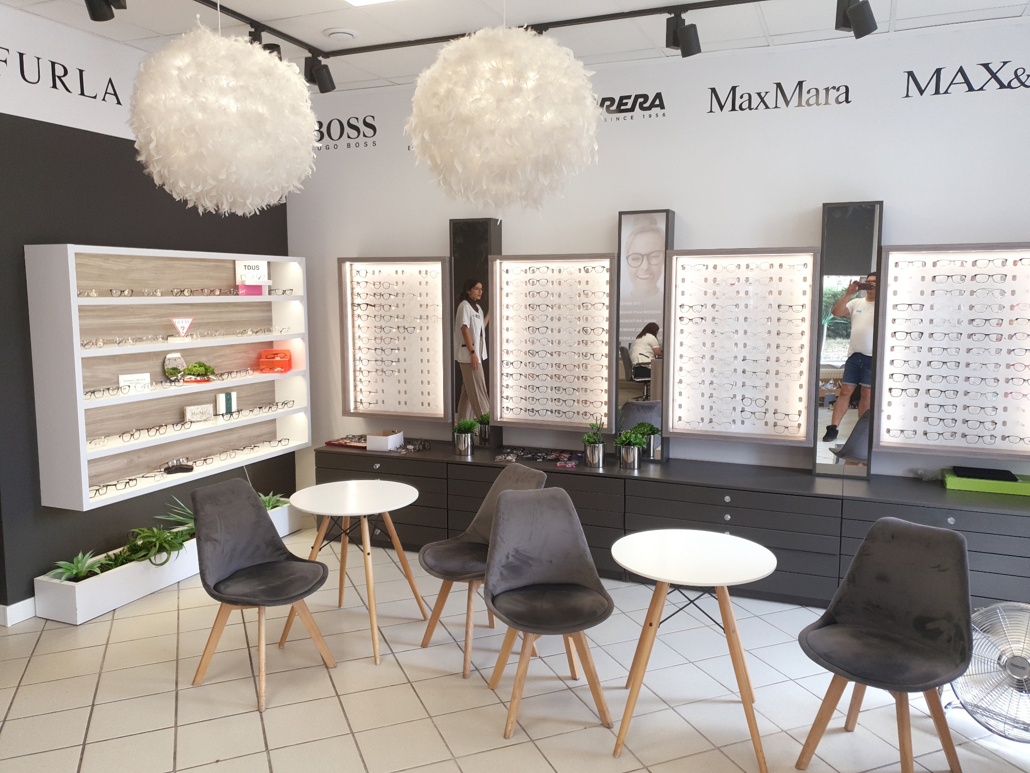 Wnętrze salonu optycznego z ekspozycją okularów w podświetlanych gablotach, szare krzesła i białe stoliki tworzą strefę dla klientów, a wiszące lampy z piór dodają elegancji.