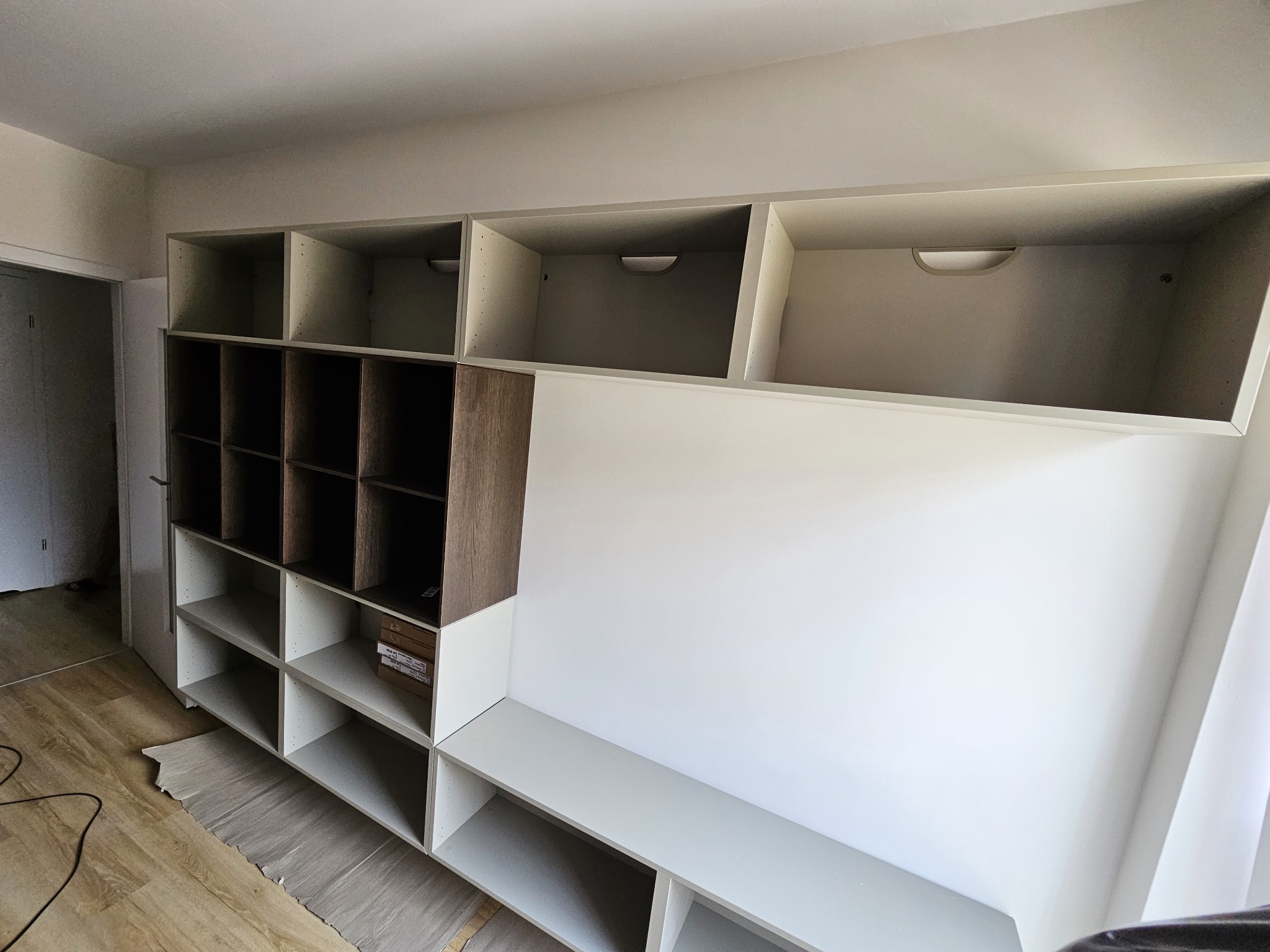 Wielopoziomowa, częściowo zamontowana biało-brązowa szafa wnękowa z otwartymi półkami, widoczne kartony na jednej z półek, zabezpieczona podłoga.