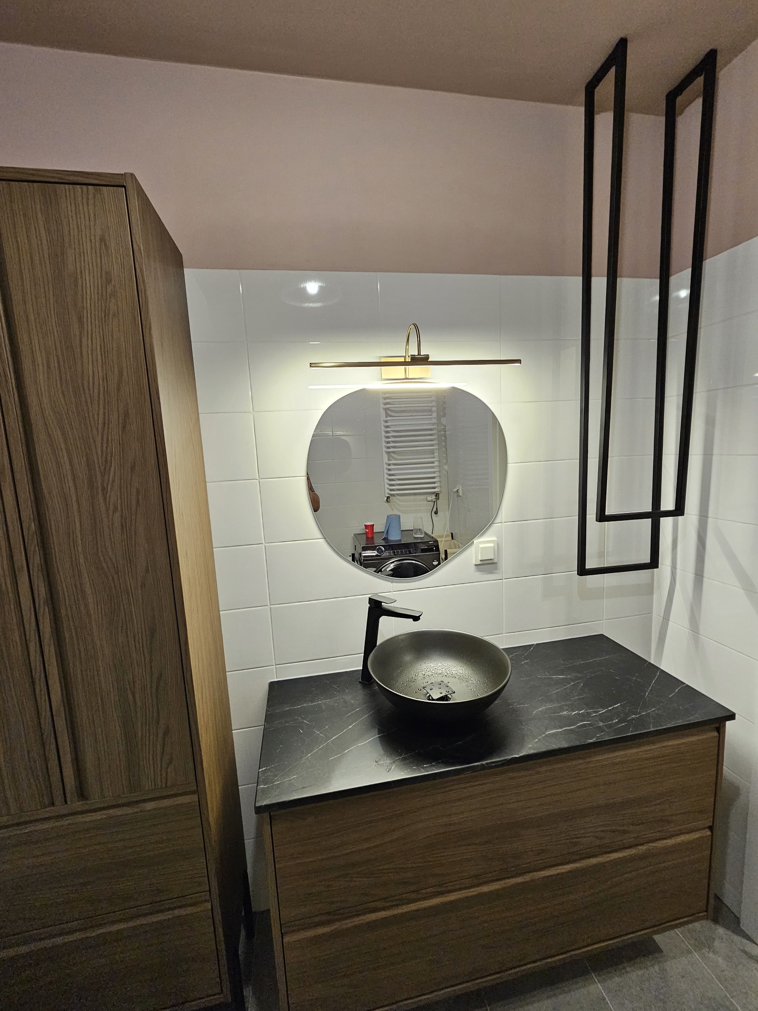 Nowoczesna łazienka z umywalką na szafce z drewnianymi frontami, okrągłym lustrem podświetlonym listwą LED i czarnymi, geometrycznymi elementami dekoracyjnymi na ścianie.