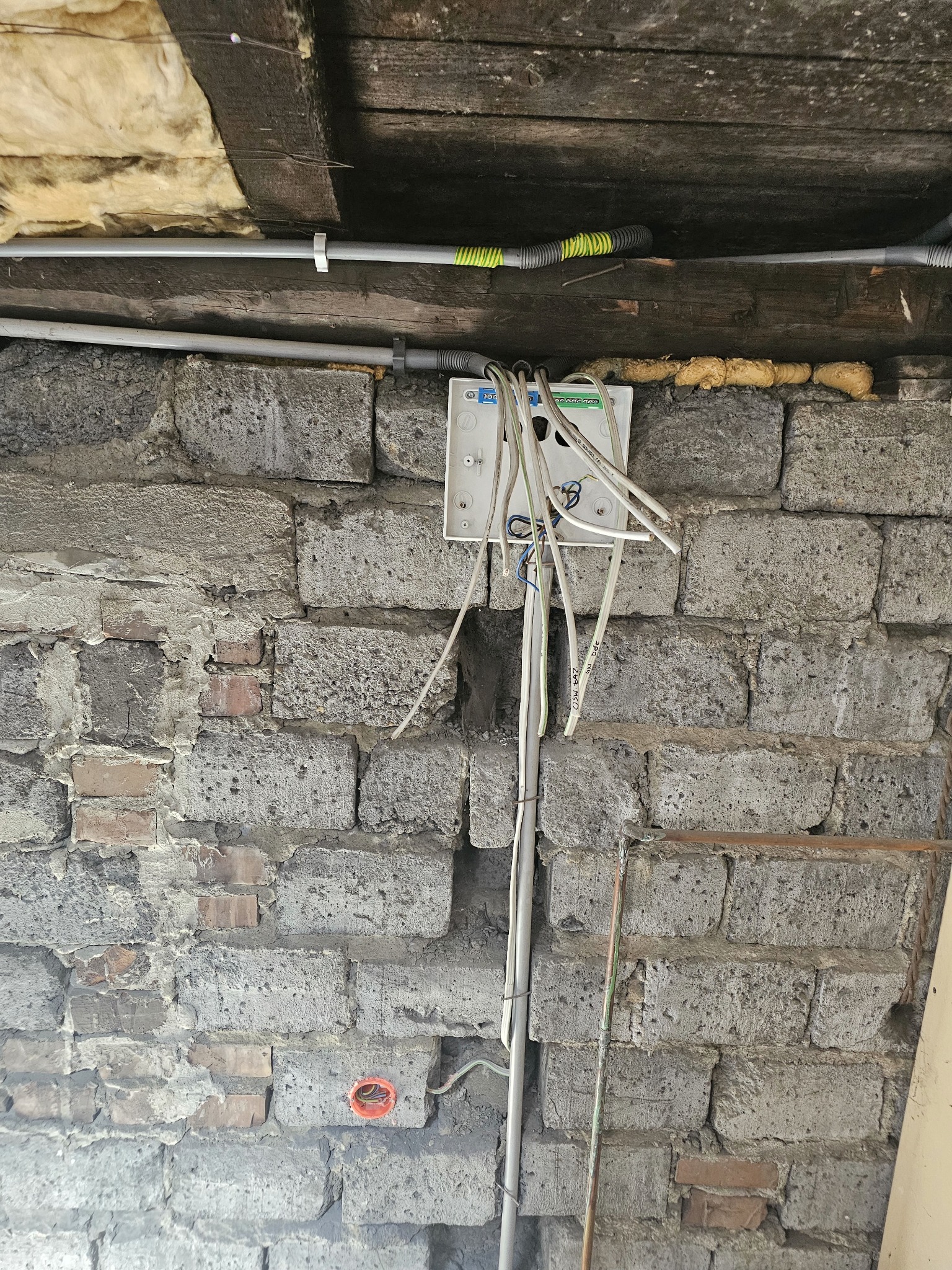 Niezabezpieczona skrzynka elektryczna z wystającymi przewodami na tle szarej, nierównej ściany z bloków betonowych i drewnianego stropu z widoczną izolacją.