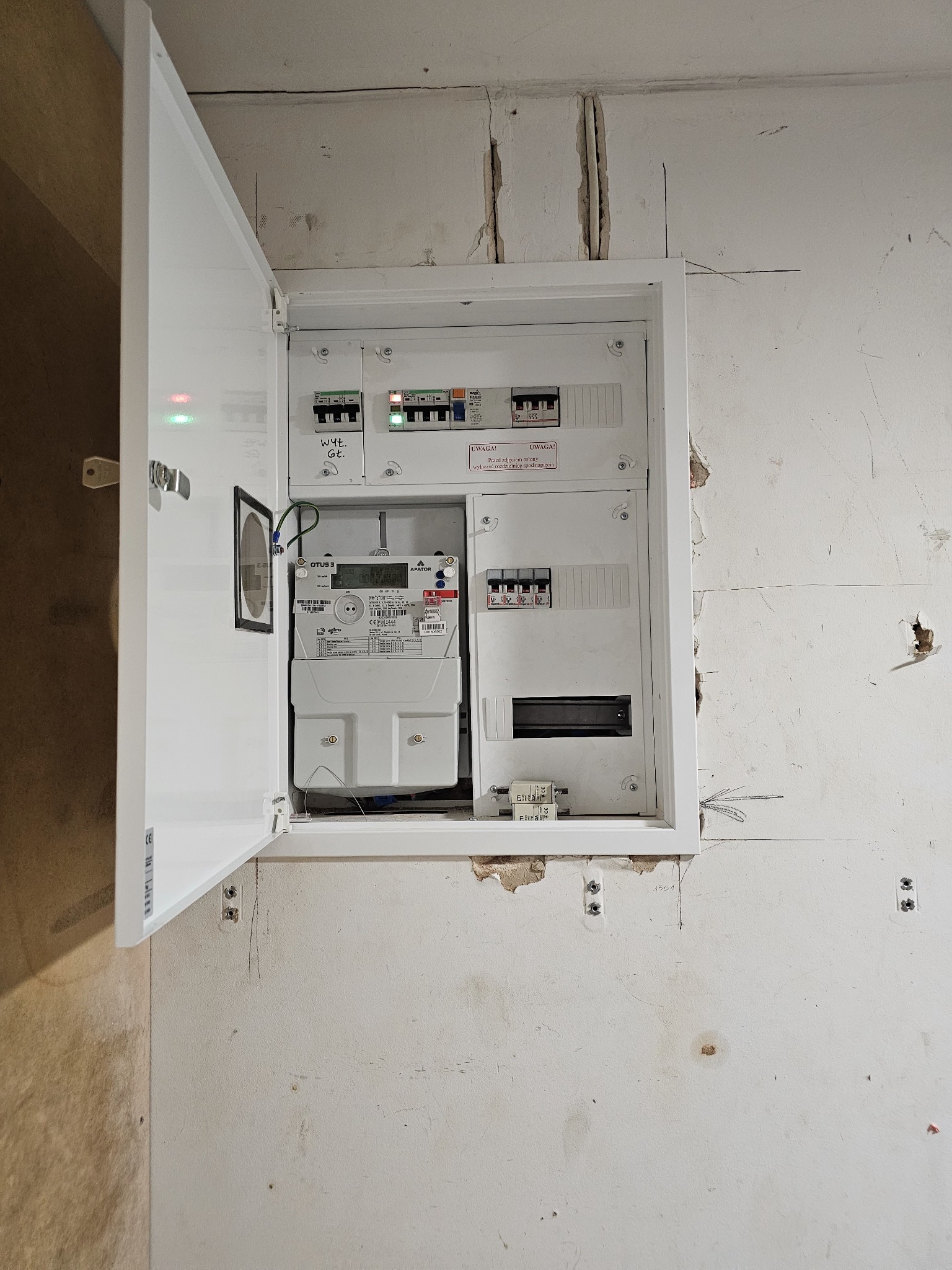 Otwarta biała skrzynka rozdzielcza z licznikiem i bezpiecznikami zamontowana na surowej, nieotynkowanej ścianie. Widoczne ślady po instalacji i rysunki na ścianie.