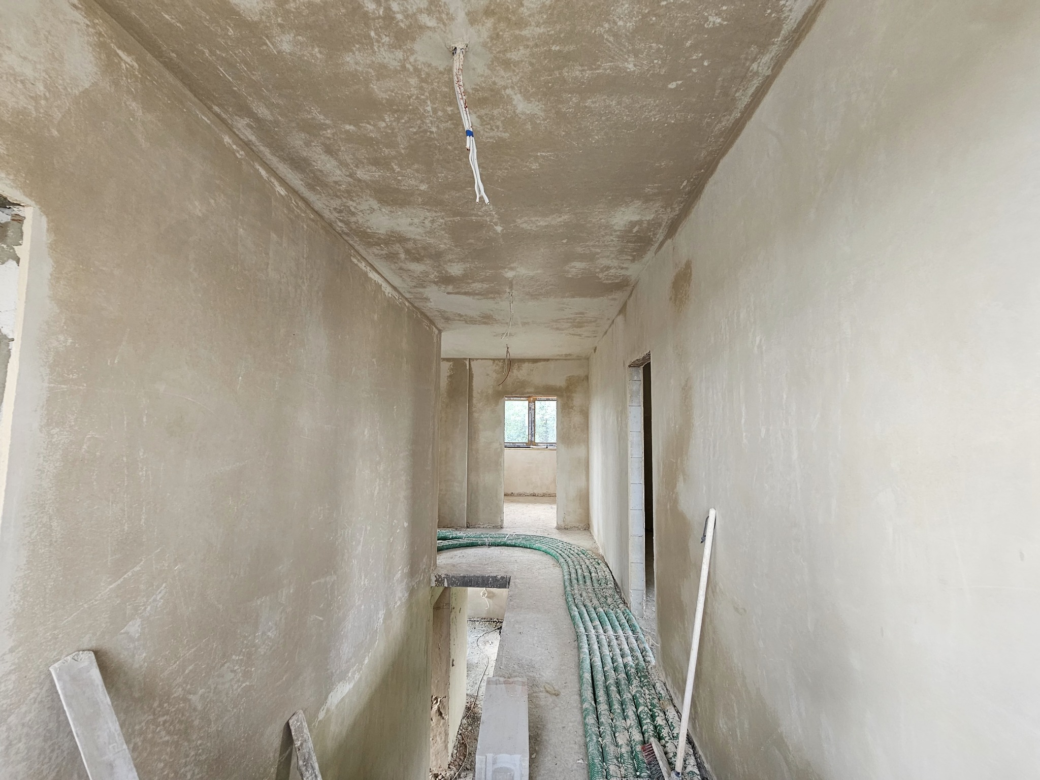 Wnętrze w stanie surowym: ściany pokryte świeżym tynkiem, widoczne przewody elektryczne na suficie i rury instalacyjne na podłodze, przygotowane do dalszych prac wykończeniowych.