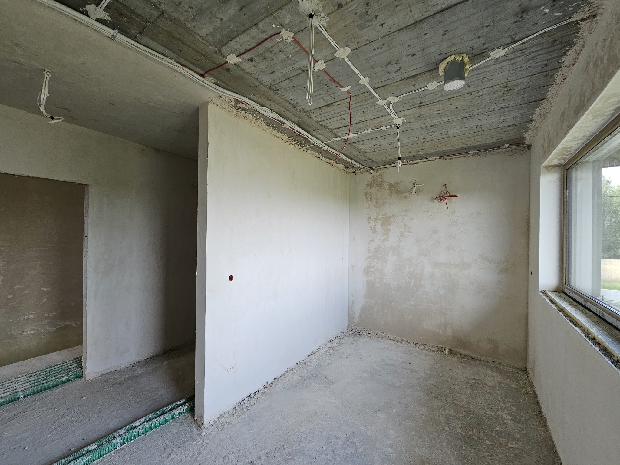 Surowy stan wnętrza z widocznymi przewodami elektrycznymi na suficie, częściowo otynkowane ściany, okno z widokiem na zieleń i fragment instalacji ogrzewania podłogowego.