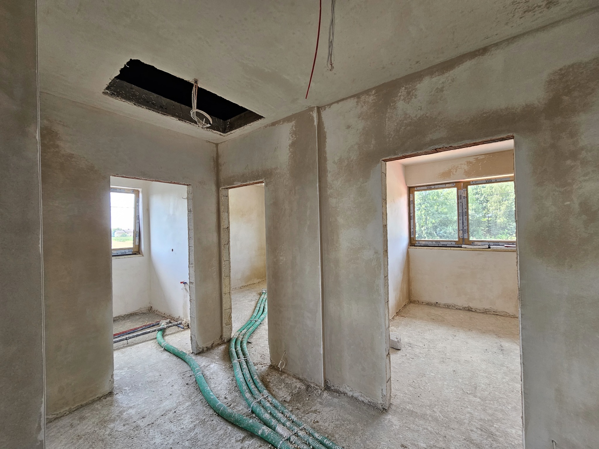 Wnętrze w Rybniku w trakcie prac tynkarskich: świeżo otynkowane ściany z widocznymi przewodami elektrycznymi i otworami w suficie, zielone węże na podłodze.