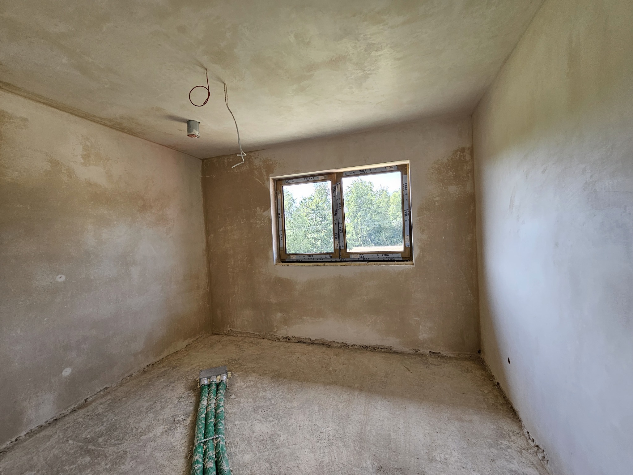 Surowe wnętrze pokoju z oknem i wstępnie nałożonym tynkiem na ścianach i suficie, widoczne wystające kable elektryczne i rury przy podłodze.