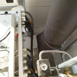 Fragment instalacji grzewczej z widocznym kotłem, rurami, zaworami i przewodami elektrycznymi w trakcie podłączenia. Widoczna izolacja termiczna na zaworze.