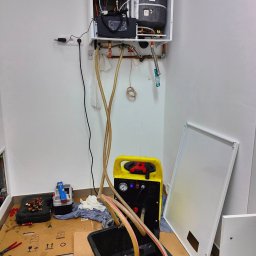 HTS ENERGY Marcin Wawrzyniak - Biały piec gazowy zamontowany na ścianie, podłączony do instalacji. Obok narzędzia i sprzęt do czyszczenia instalacji. Widoczne rury i kable.