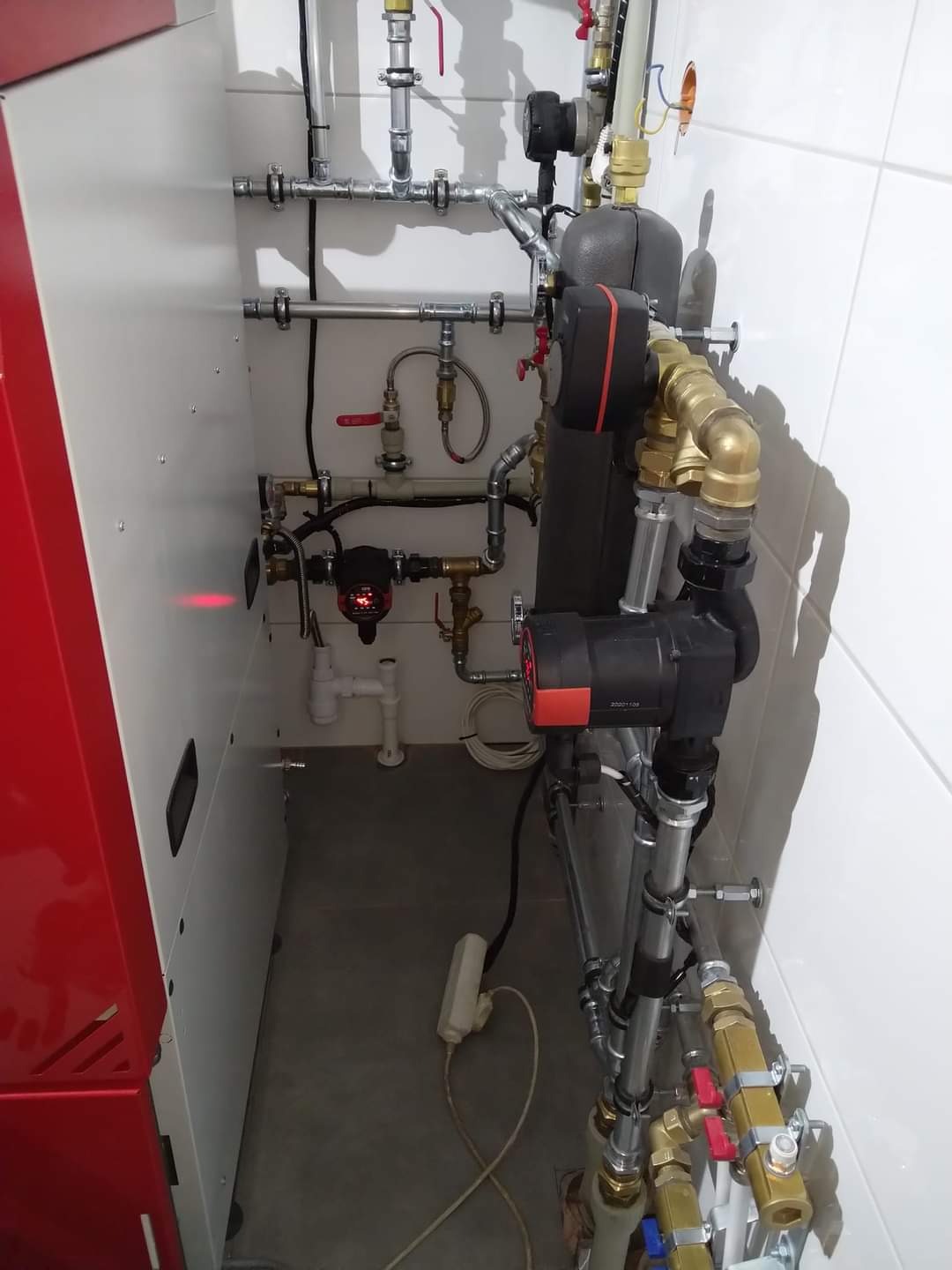 Złożona instalacja grzewcza z kotłem z czerwonym akcentem, rurami miedzianymi i stalowymi, pompą obiegową i białą ceramiką w tle. Widoczny kabel zasilający leżący na podłodze.