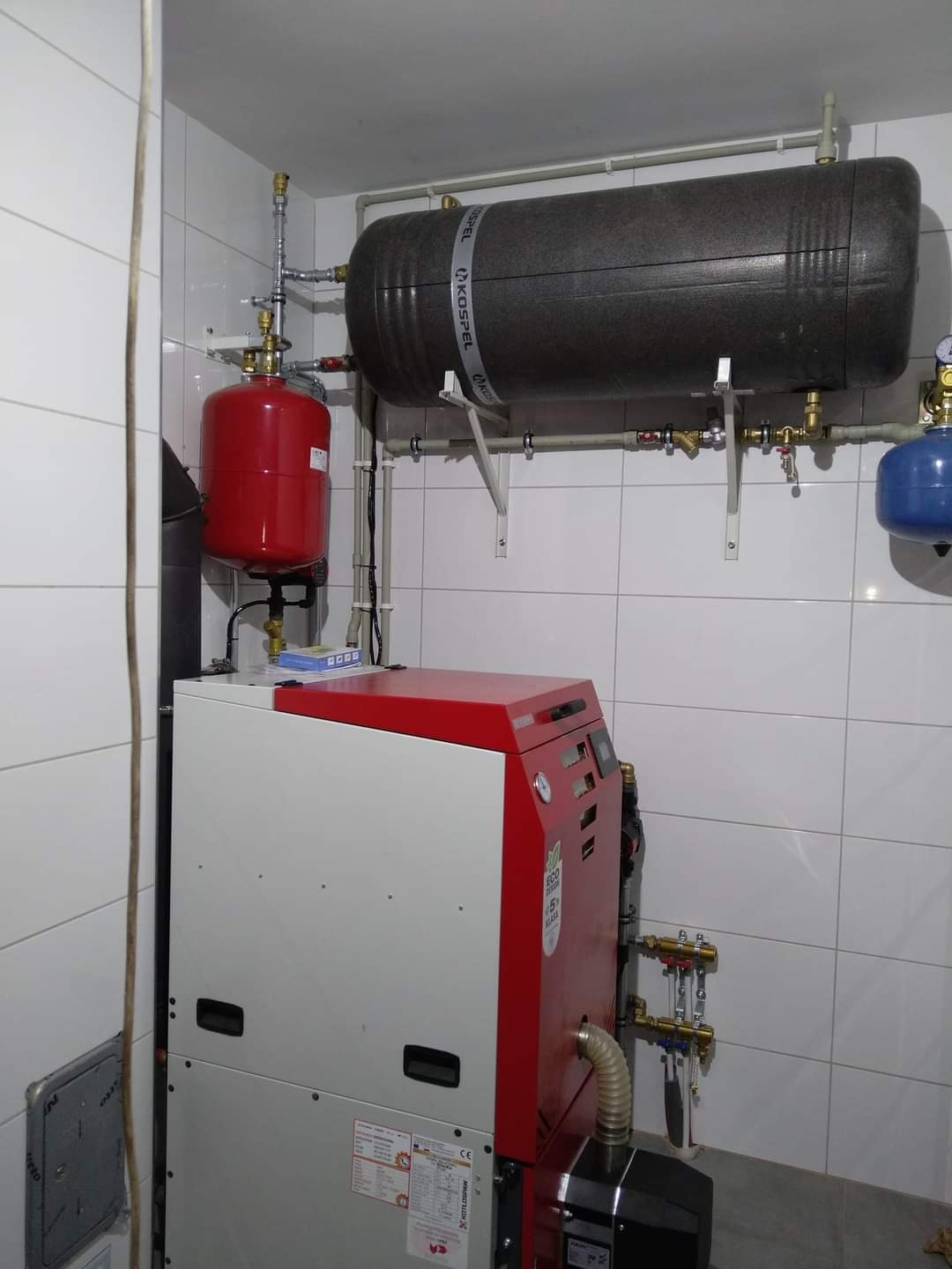 Zestaw urządzeń grzewczych Kospel w kotłowni z białymi płytkami: piec z czerwoną obudową, czerwony zbiornik wyrównawczy i szary zasobnik ciepłej wody na ścianie.